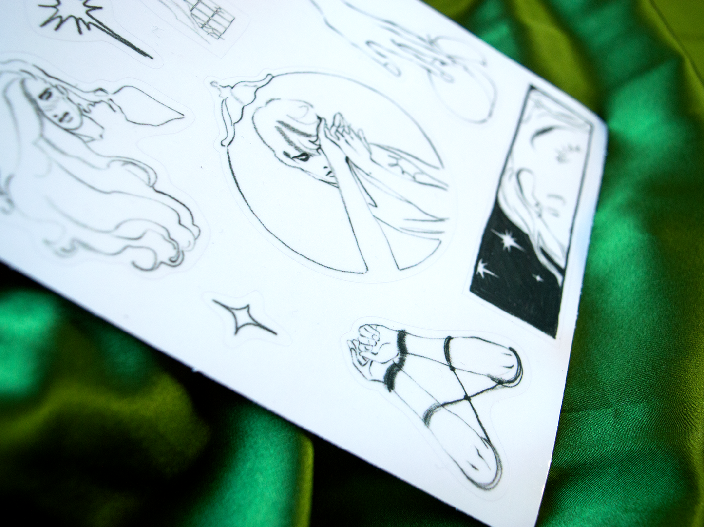 The transparent sticker sheet of a collection of small pencil illustrations from the Kinktober zine, close-up.
